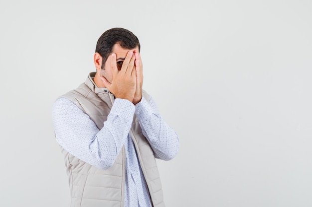 Young man in beige jacket covering face with hands and looking through fingers and looking optimistic , front view.