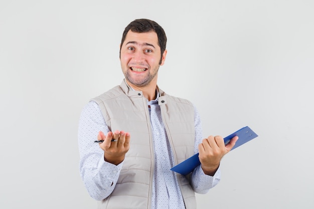 Young man in beige jacket raising hand in questioning manner while holding notebook and pen and looking optimistic , front view.