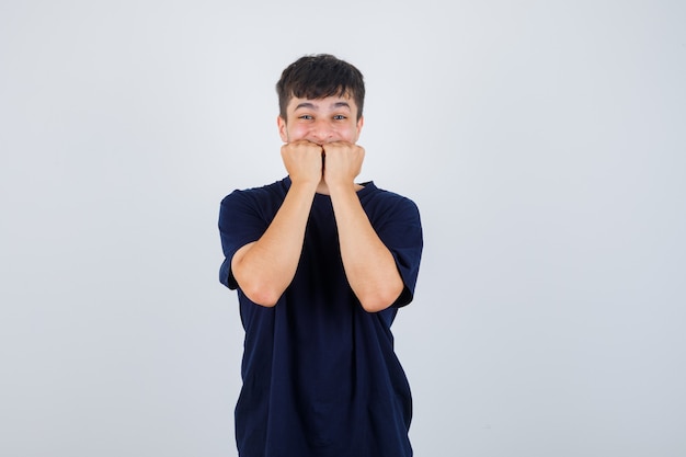 Young man biting fists emotionally in black t-shirt and looking scared. front view.