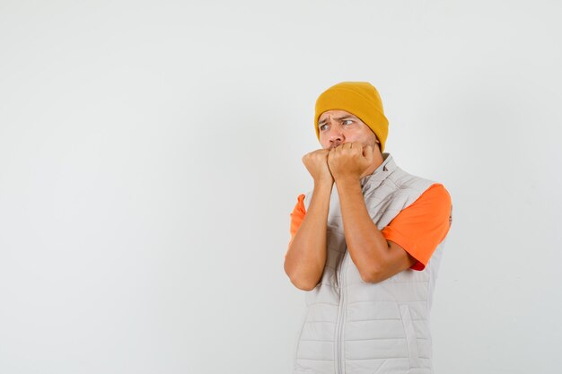 Young man biting fists emotionally in t-shirt, jacket, hat and looking scared , front view.