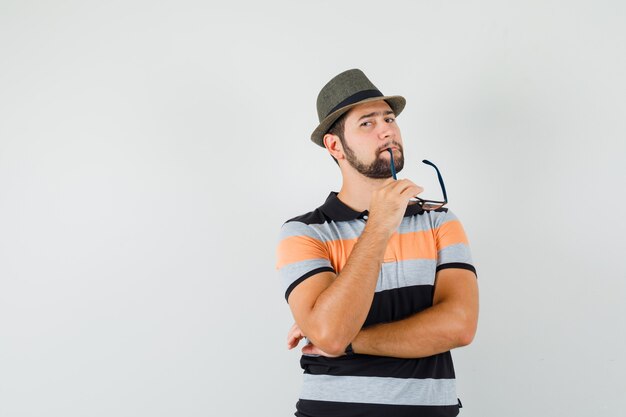 Young man biting glasses in t-shirt, hat and looking thoughtful , front view.