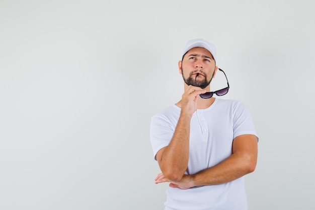 Young man biting glasses while looking up in white t-shirt and looking pensive. front view. space for text