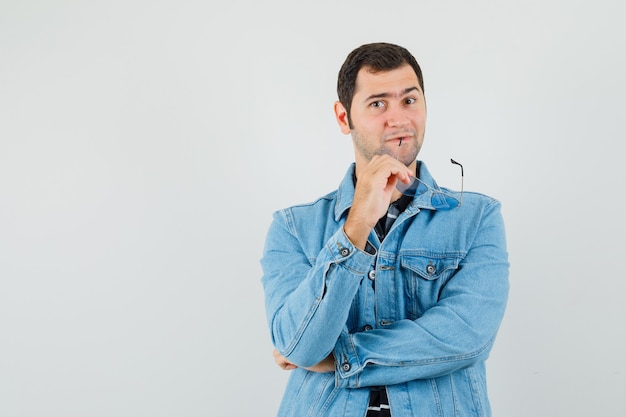 Young man biting glasses while thinking in t-shirt, jacket and looking confident. 