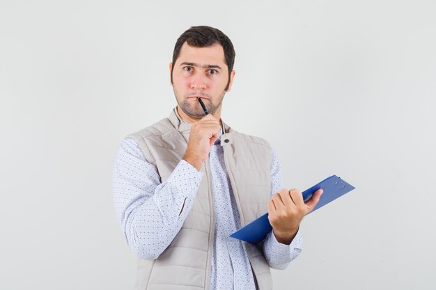 Young man biting pen while holding notebook in beige jacket and looking pensive. front view.