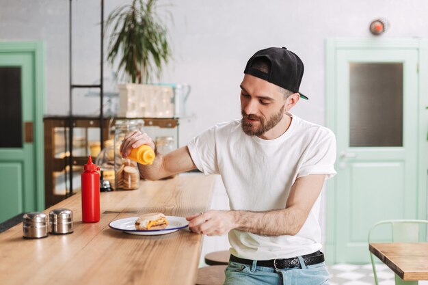 Young man in black cap sitting at the bar counter and holding mustard bottle while eating burger in cafe