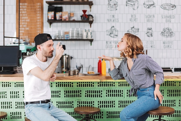 Молодой человек в черной кепке сидит за барной стойкой и фотографирует красивую даму, которая посылает ему воздушный поцелуй, проводя время вместе в кафе
