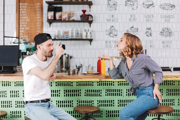 Young man in black cap sitting at the bar counter and taking photos of beautiful lady which sending him air kiss while spending time in cafe together