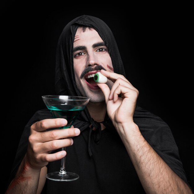 Young man in black cloak posing in studio with artificial eye and beverage