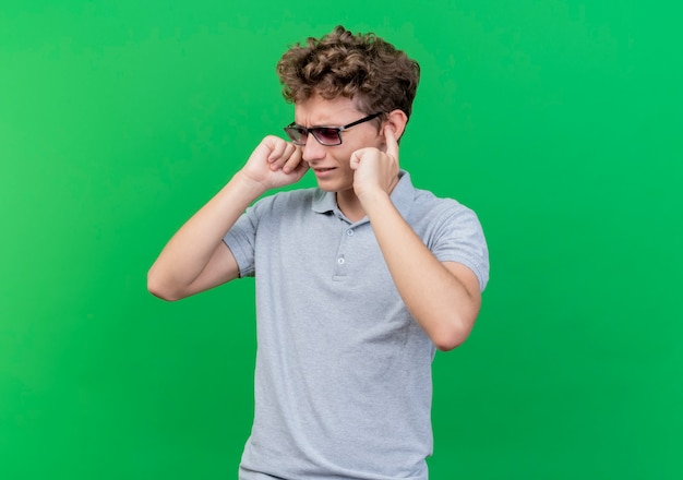 Young man in black glasses wearing grey polo shirt closing ears with fingers with annoyed expression standing over green wall