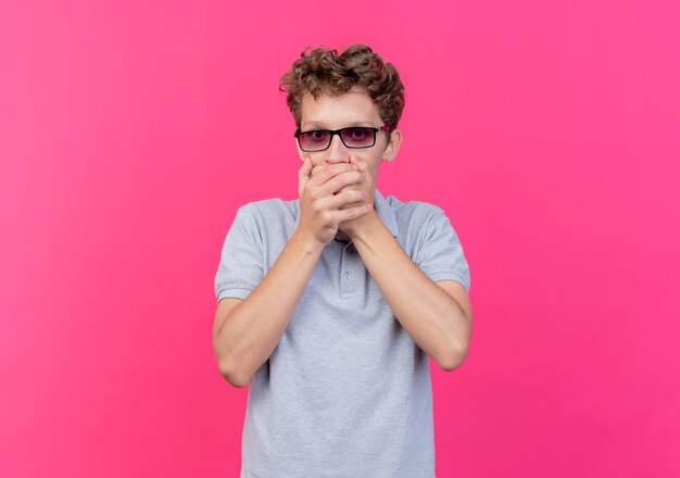 Young man in black glasses wearing grey polo shirt covering mouth with hands being shocked over pink