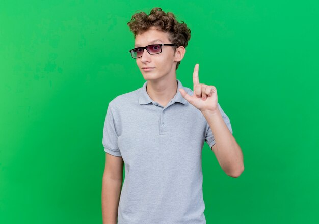Young man in black glasses wearing grey polo shirt  happy and positive showing index finger having new idea standing over green wall