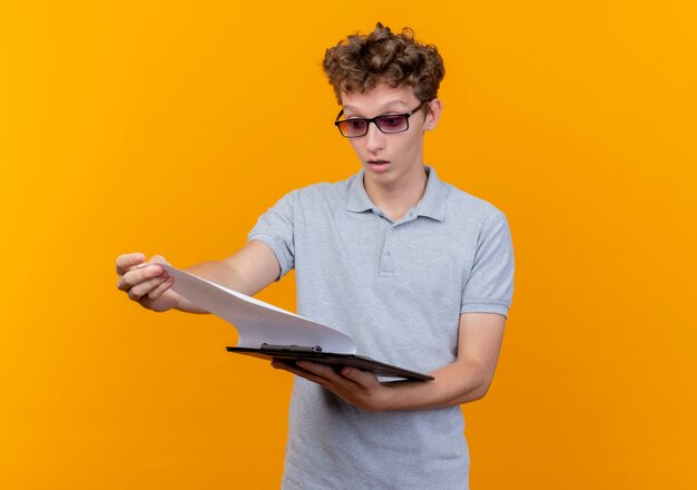 Young man in black glasses wearing grey polo shirt holding clipboard with blank pages looking at it surprised over orange