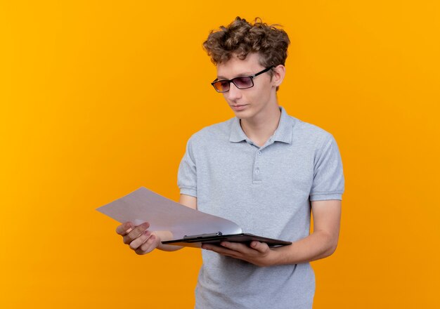 Young man in black glasses wearing grey polo shirt holding clipboard with blank pages looking at it with serious face standing over orange wall