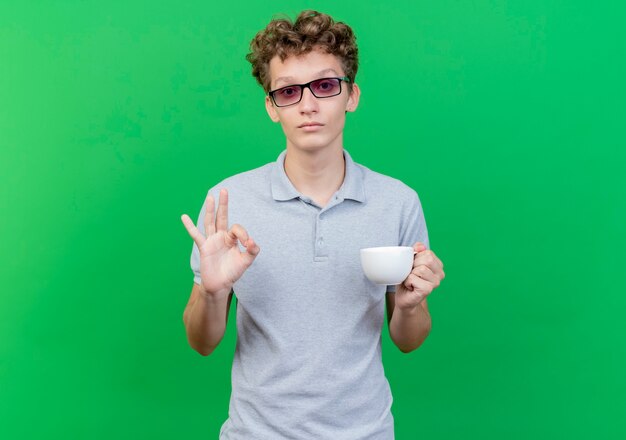 Young man in black glasses wearing grey polo shirt holding coffee cup  happy and positive showing ok sign standing over green wall