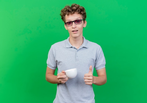Young man in black glasses wearing grey polo shirt holding coffee cup  smiling showing thumbs up standing over green wall