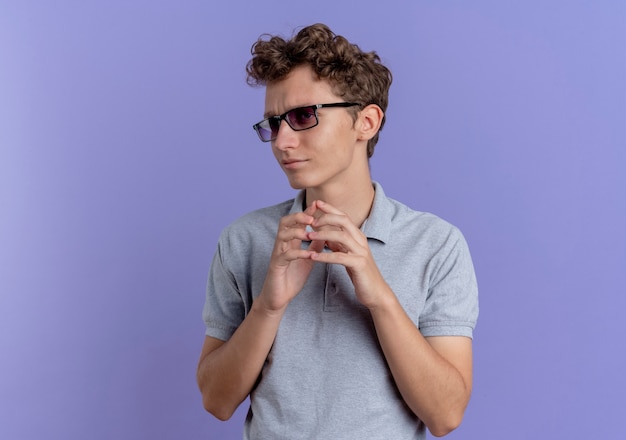 Young man in black glasses wearing grey polo shirt holding hands together looking aside displeased standing over blue wall