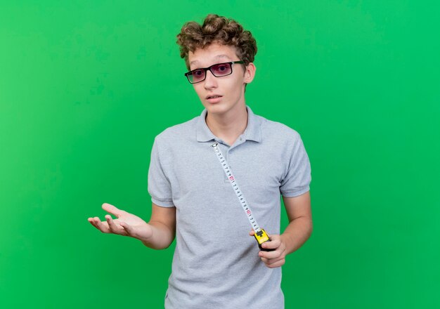 Free photo young man in black glasses wearing grey polo shirt holding measure tape gesturing with hand being confused standing over green wall