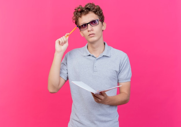 Young man in black glasses wearing grey polo shirt holding notebook with pen looking with pensive expression thinking standing over pink wall