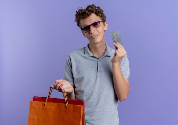 Foto gratuita giovane uomo con gli occhiali neri che indossa una polo grigia che tiene i sacchetti di carta e la carta di credito che sorride in piedi sopra la parete blu