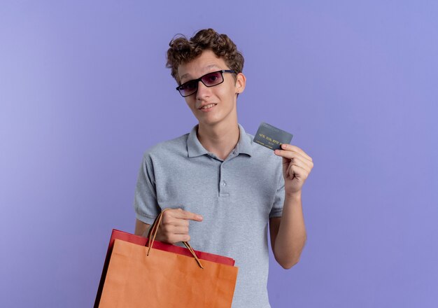 파란색 벽 위에 서 웃는 신용 카드를 보여주는 종이 가방을 들고 회색 폴로 셔츠를 입고 검은 안경에 젊은 남자