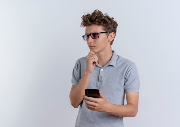 Young man in black glasses wearing grey polo shirt holding smartphone looking aside with hand on chin with pensive expression standing over white wall