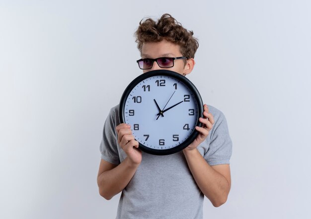 Young man in black glasses wearing grey polo shirt holding wall clock hiding his face behind it standing over white wall