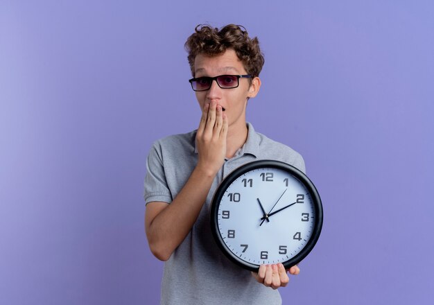 Young man in black glasses wearing grey polo shirt holding wall clock looking surprised and amazed standing over blue wall