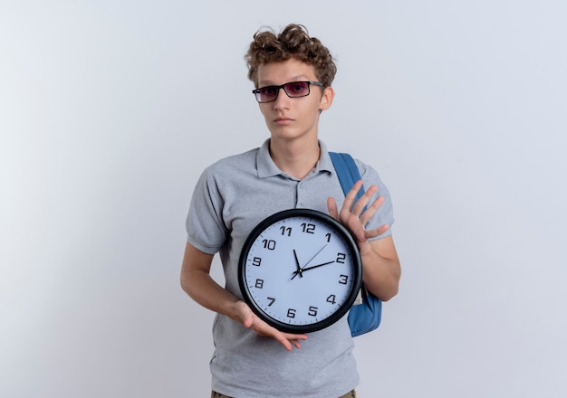 Young man in black glasses wearing grey polo shirt holding wall clock  with serious face standing over white wall