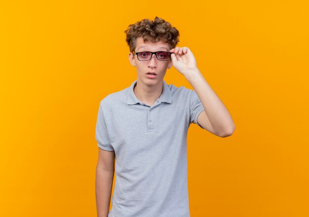 Young man in black glasses wearing grey polo shirt lookign at camera concentrating hard touching his glasses over orange