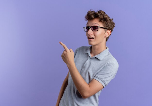 Young man in black glasses wearing grey polo shirt looking aside happy and surprised pointing with finger to the side over blue