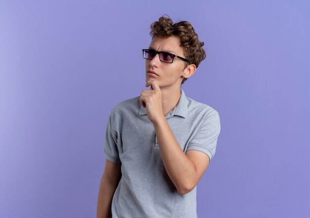 Молодой человек в черных очках, одетый в серую рубашку поло, озадаченно смотрит в сторону, стоя у синей стены