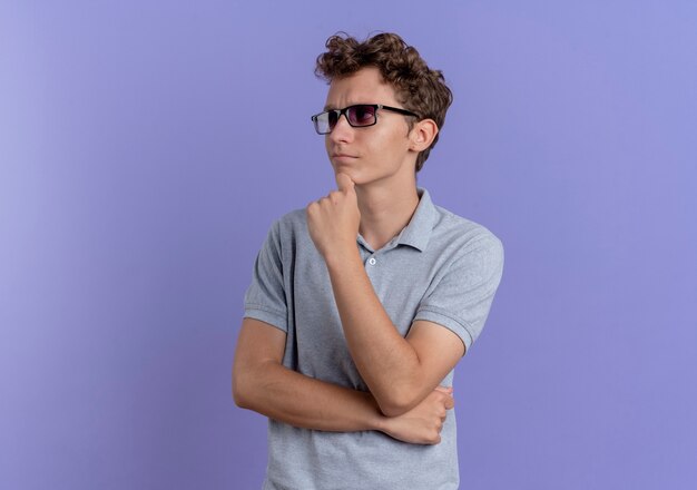 Young man in black glasses wearing grey polo shirt looking aside with pensive expression over blue