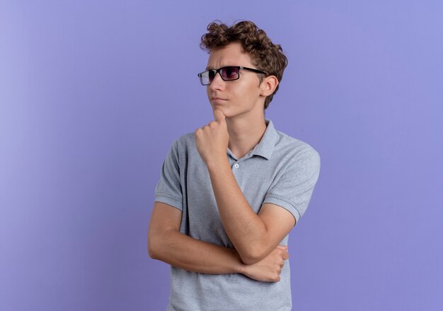 Молодой человек в черных очках в серой рубашке поло смотрит в сторону с задумчивым выражением лица поверх синего