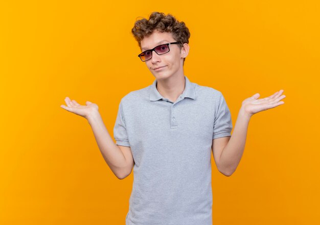 Young man in black glasses wearing grey polo shirt looking confused and uncertain raising hands having no answer over orange