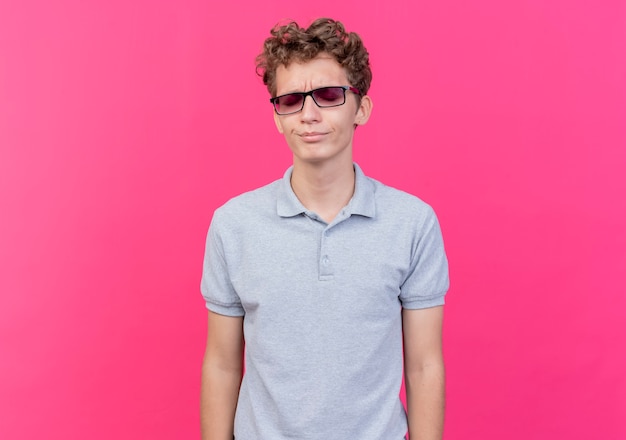 Young man in black glasses wearing grey polo shirt looking displeased standing over pink wall