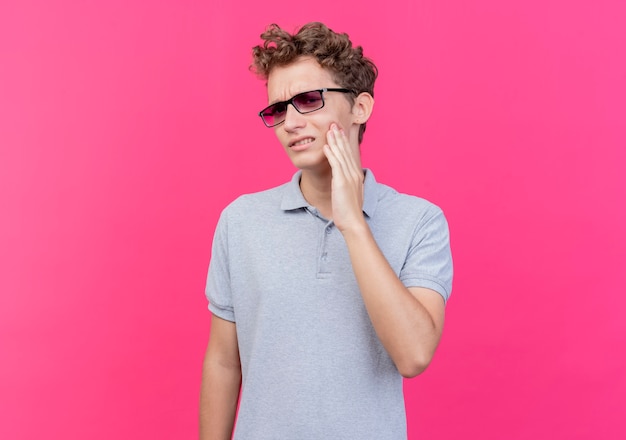 Young man in black glasses wearing grey polo shirt looking unwell touching his cheek having toothache standing over pink wall