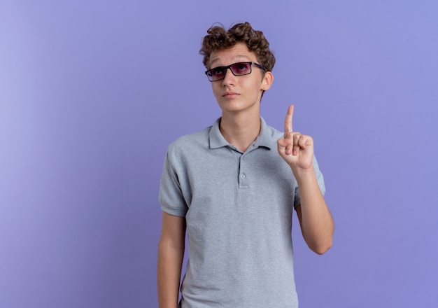 Young man in black glasses wearing grey polo shirt looking up pointign with index finger at something standing over blue wall