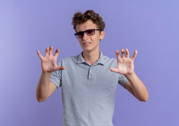 Young man in black glasses wearing grey polo shirt making defense gesture with disgusted expression standing over blue wall