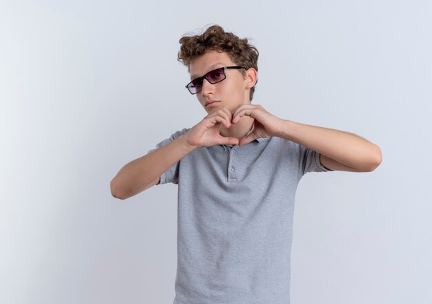 Young man in black glasses wearing grey polo shirt  making heart gesture with fingers standing over white wall