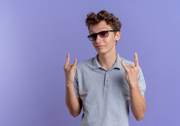 Young man in black glasses wearing grey polo shirt  making rock symbols smiling standing over blue wall