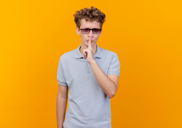 Молодой человек в черных очках в серой рубашке поло делает жест молчания с пальцем на губах, стоя над оранжевой стеной