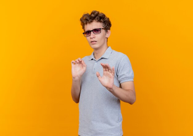 Young man in black glasses wearing grey polo shirt making stop sign holding hands out as telling do not come closer scared standing over orange wall