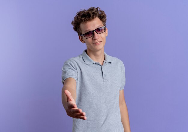 Young man in black glasses wearing grey polo shirt offering hand smiling frinedly greeting gesture standing over blue wall