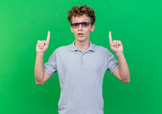 녹색 벽 위에 서서 놀란 idex 손가락으로 가리키는 회색 폴로 셔츠를 입고 검은 안경에 젊은 남자
