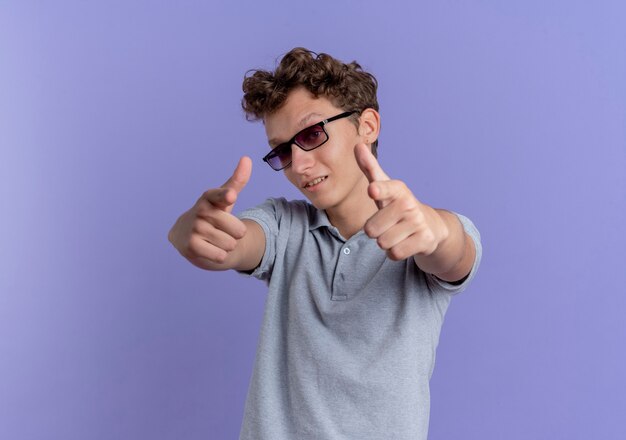 Young man in black glasses wearing grey polo shirt pointing with index fingers  looking confident standing over blue wall