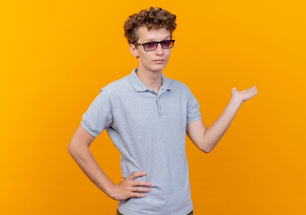 Young man in black glasses wearing grey polo shirt presenting something with arm of hand looking confident over orange