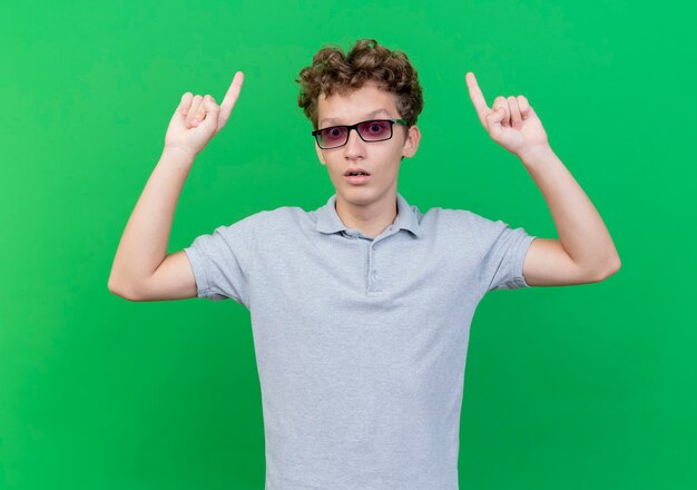 Young man in black glasses wearing grey polo shirt raising both hands showing index fingers being surprised standing over green wall