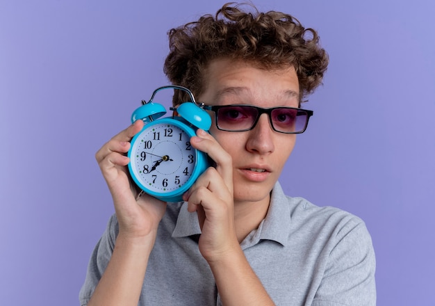 Young man in black glasses wearing grey polo shirt showing alarm clock  being confused standing over blue wall