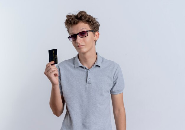 Young man in black glasses wearing grey polo shirt showing credit card looking confident standing over white wall