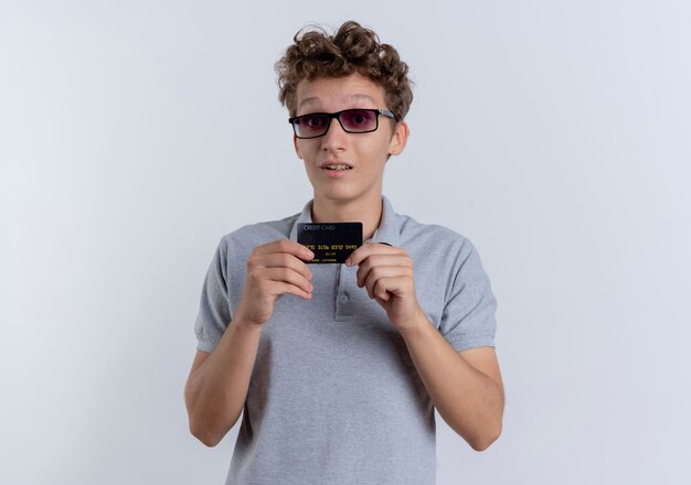 Young man in black glasses wearing grey polo shirt showing credit card  surprised standing over white wall